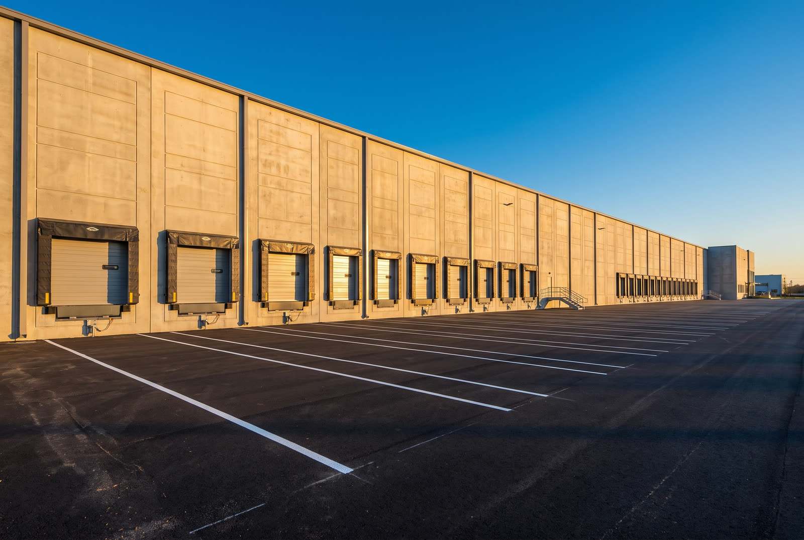 Newly completed tilt-up concrete distribution centre with a long row of loading docks, a paved truck court, and two workers in hard hats and high-visibility vests walking the dock apron